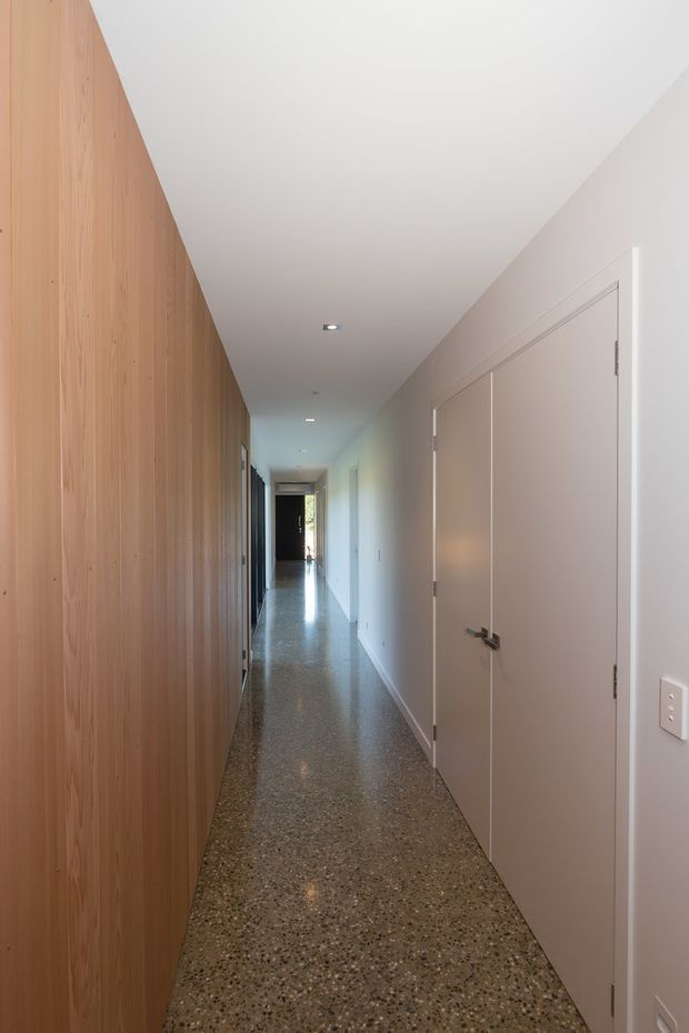 A long, narrow contemporary hallway with a full-height light timber feature wall and concealed storage or doors along the left side. The opposite wall is painted a soft warm white with flush-panel doors, while the polished exposed aggregate or terrazzo-like floor reflects recessed ceiling lights. The corridor leads toward a darker doorway at the far end and has a clean, minimal architectural character.