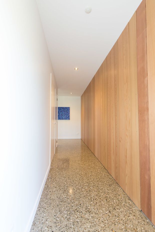 A long, narrow minimalist hallway with a white plaster wall on one side and full-height vertical timber wall panelling with concealed doors or joinery on the other. The floor is finished in speckled polished terrazzo, with recessed ceiling lights and a small blue artwork at the far end.