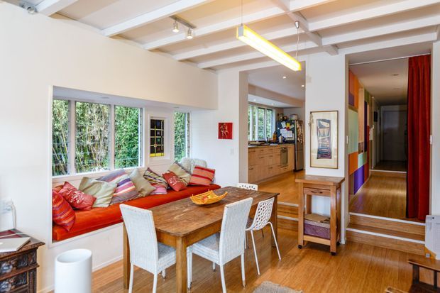 Bright open-plan dining and living area with a built-in window seat, colourful cushions, a rustic timber dining table, white woven dining chairs, and warm timber flooring. The room connects visually to a compact timber kitchen and a hallway with bold multicoloured wall panels and red curtains. Exposed painted ceiling beams and large windows contribute to an eclectic, characterful interior.