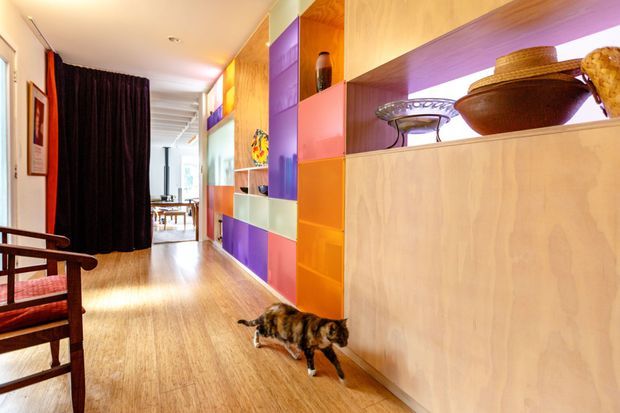 A colourful, contemporary interior hallway with warm timber flooring and extensive custom built-in cabinetry. The cabinetry combines natural timber panels with vivid purple, orange, pink, blue and green recessed sections, some softly illuminated for display. A dark curtain separates the corridor from a brighter dining or living area beyond. A timber-framed chair sits at the left, while decorative objects are displayed in open shelving on the right.