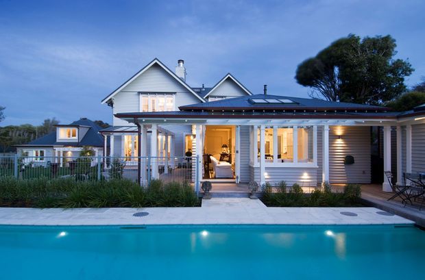 Dusk exterior view of a renovated light-coloured weatherboard home beside a swimming pool. The house combines a low-pitched dark roofed extension with gabled roof forms, white trim, a covered verandah, illuminated windows, and landscaped planting around the pool.