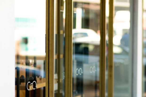 Close-up view of a glazed commercial storefront or entrance with tall vertical glass panels and slim brass-toned metal frames. Reflections and blurred views of the street and interior are visible through the glazing, with small white lettering on the glass. The image is shallow-focus and abstract, emphasizing the repeated framed doors or windows rather than a clearly identifiable room.