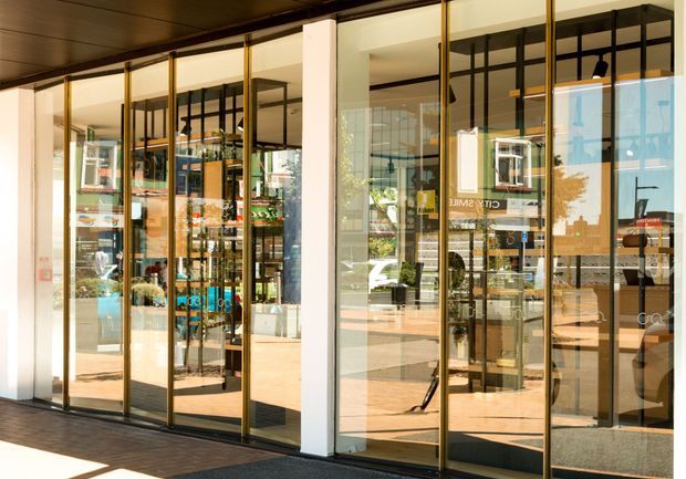 Street-facing commercial shopfront with extensive floor-to-ceiling glazed folding or framed doors, slim dark metal and gold-toned framing, white structural piers, and a dark soffit. Shelving, retail displays, and a customer-facing interior are visible through the glazing, with strong reflections of the surrounding street.