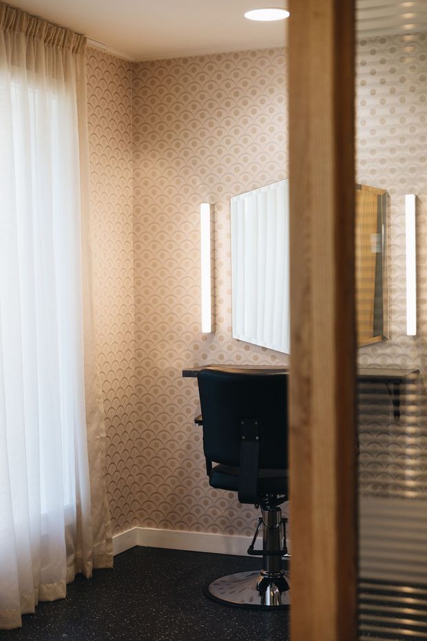 A compact, warmly lit salon or beauty treatment room viewed through a partially open timber-framed glass door. The room has patterned scallop wallpaper, sheer curtains over a tall window, a black salon chair, illuminated wall mirror, and dark speckled flooring. The reflected view shows additional salon fixtures and joinery.
