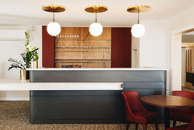 A refined hospitality bar or reception counter with dark charcoal built-in cabinetry, a pale stone or solid-surface countertop, warm timber shelving displaying glassware, and deep red wall panels. Three spherical pendant lights with brass-toned ceiling plates hang above the counter. Red upholstered chairs and a round timber table sit in the foreground, while patterned carpet and white walls complete the space.