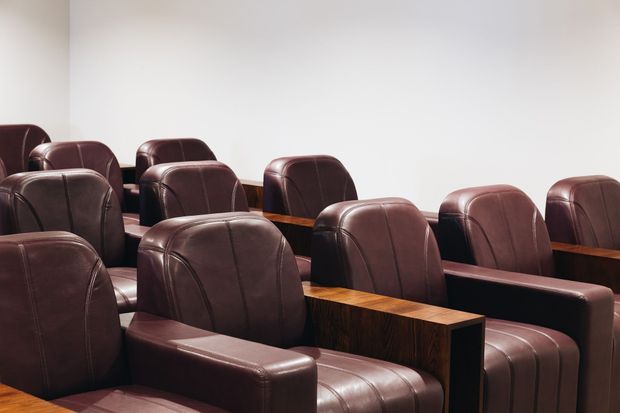 Rows of dark brown leather upholstered theatre or auditorium seats with broad wooden armrests and dividers, arranged in a compact interior against plain white walls.