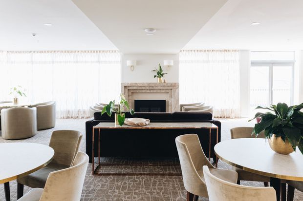 Bright, refined open-plan living and dining room with large sheer-curtained windows, a central stone fireplace, dark console cabinetry, round dining tables, upholstered chairs, and a neutral patterned rug. The space has a warm contemporary-classic feel with abundant natural light.