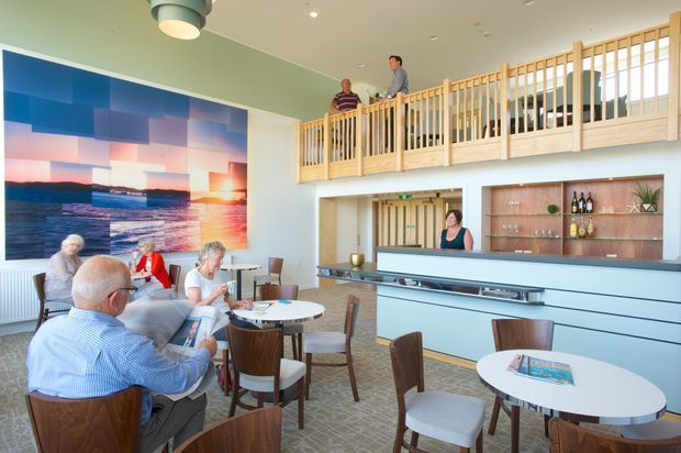 A bright, contemporary hospitality lounge or café with a double-height interior and mezzanine-level timber balustrade. The space includes a long pale reception or bar counter, built-in timber shelving with bottles and decor, round café tables, upholstered chairs, and a large ocean-themed wall artwork. Several people are seated and standing in the space.