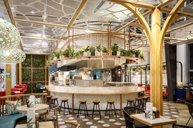 A large, stylish hospitality interior centred on a curved island bar and service counter. The space features extensive light timber branching columns and arches, a patterned geometric ceiling, hanging sculptural pendant lights, abundant potted greenery, patterned tiled flooring, and varied café seating including stools, booths, and tables. The open plan includes a mezzanine-like upper level and a visible commercial kitchen/service area.