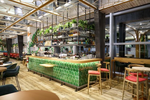 A bright, open-plan hospitality interior centered on a long green tiled bar counter. The bar has a timber countertop, decorative bottle shelving, overhead planting, stools with red upholstered seats, and surrounding café or restaurant tables and chairs. Exposed structural framing, industrial-style glazing, timber flooring, and a high ceiling create a lively contemporary venue atmosphere.