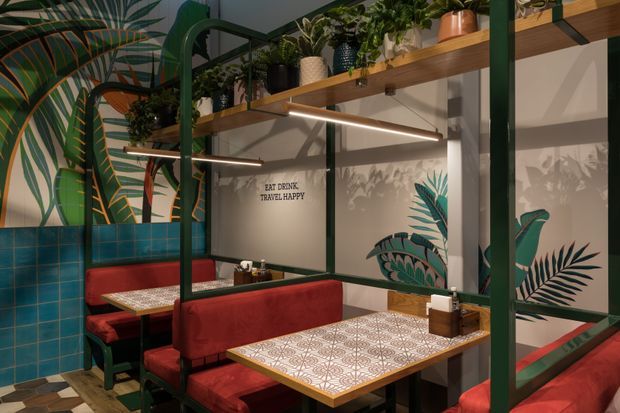 A compact, richly decorated commercial dining space with red upholstered booth seating, patterned tiled tabletops, teal wall tiles, dark green metal framing, and abundant tropical botanical graphics. Timber shelves with potted plants run along the upper wall, while warm linear lighting and large mirrored or glazed panels create a layered, intimate atmosphere.