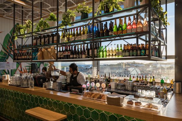 A contemporary café or bar interior with a long timber counter, green geometric tiled front, coffee equipment, beer taps, pastries, and an extensive suspended black metal shelving display stocked with bottles and plants. Large windows overlook a waterfront or harbour-side urban setting, while staff work behind the counter.