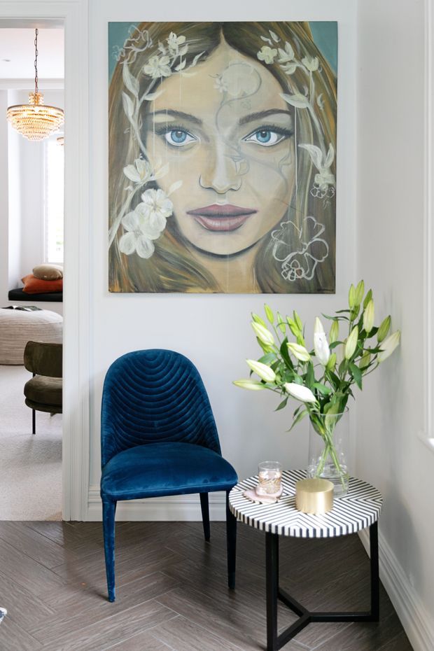 A stylish interior vignette in a light-painted hallway or entry area, featuring a vivid deep-blue upholstered accent chair beside a small black side table with a bold black-and-white striped top. A vase of white lilies, a candle, and a large floral portrait artwork add decorative character. Through the open doorway, part of a bedroom with a bed and pendant light is visible. Dark herringbone timber flooring and white architectural trim frame the scene.