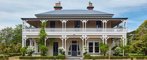 Front exterior view of a two-storey historic-style residence with a broad hipped metal roof, twin chimneys, extensive wraparound verandahs, ornate white timber fretwork and balustrades, pale weatherboard cladding, and a centrally positioned dark entrance door. Established garden planting frames the facade.