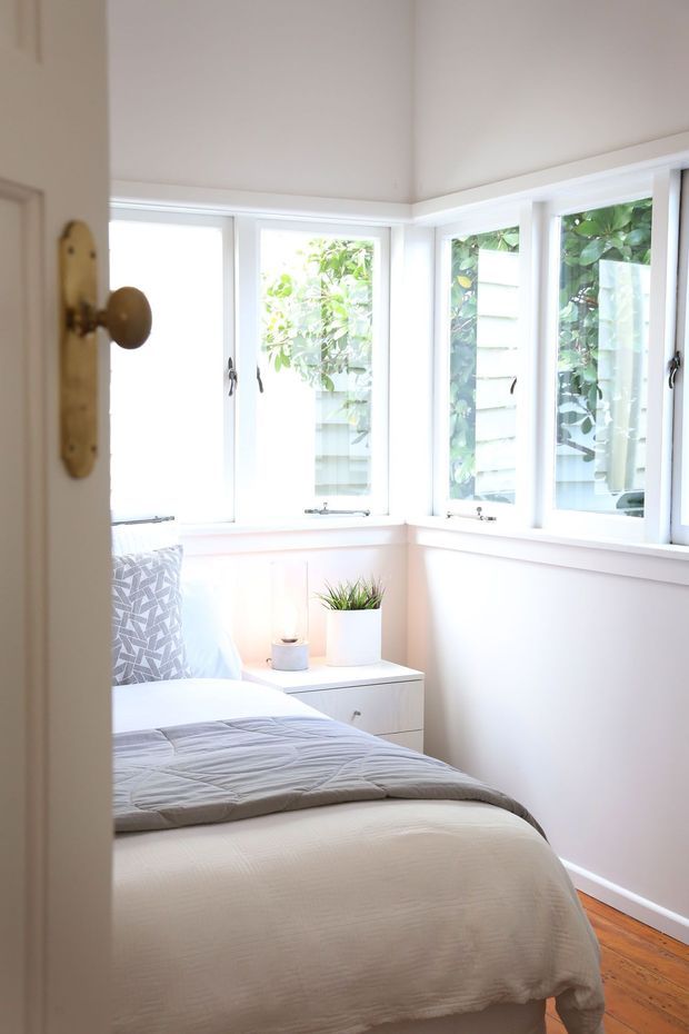 A bright, compact bedroom viewed through a partially open door. The room has a bed with pale bedding, a small white bedside cabinet, potted plants, white walls, extensive corner windows with white frames, and warm timber flooring. Lush greenery is visible outside the windows. The styling is simple, fresh, and understated.