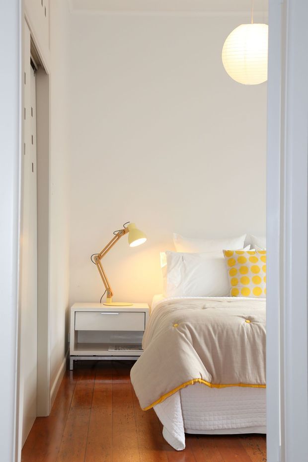A bright, compact bedroom viewed through an open doorway. The room has white walls and bedding, warm timber floorboards, a simple bedside table with a yellow articulated lamp, a yellow-and-white polka-dot cushion, and a spherical pendant light. The styling is clean, playful, and contemporary with a warm colour accent.