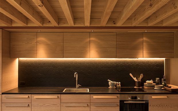Warm, sophisticated contemporary kitchen with extensive timber cabinetry and exposed timber ceiling beams. A dark hexagonal tile splashback is dramatically illuminated by concealed under-cabinet lighting, contrasting with the timber surfaces. The kitchen includes a long benchtop with integrated sink, cooktop and built-in oven, with carefully arranged utensils and crockery.