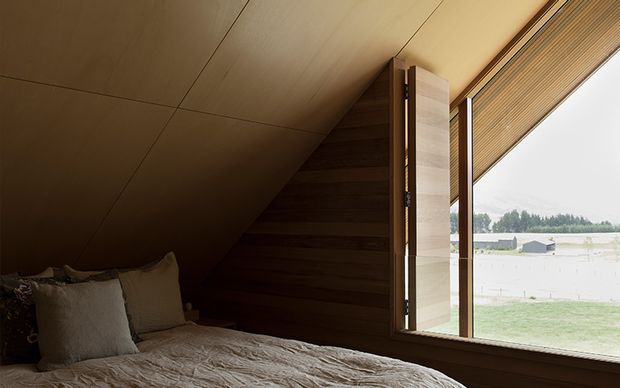 A compact bedroom beneath a steeply sloping timber-lined roof, with a low bed and neutral bedding in the foreground. Dark horizontal timber wall panelling forms the headboard area, while a large angled window with a timber blind provides views across a rural landscape. The room has a warm, intimate, cabin-like atmosphere and restrained natural materials.