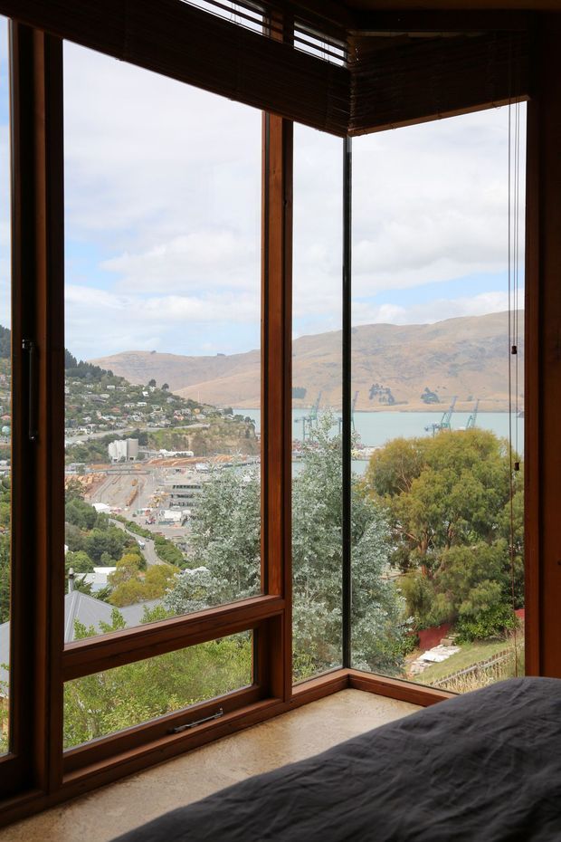 A bedroom corner with expansive floor-to-ceiling corner glazing overlooking a lake, hillside settlement, and surrounding mountains. Warm timber window frames and woven timber blinds frame the view, while a dark bed occupies the foreground. The room has a calm, nature-connected atmosphere and strong natural outlook.