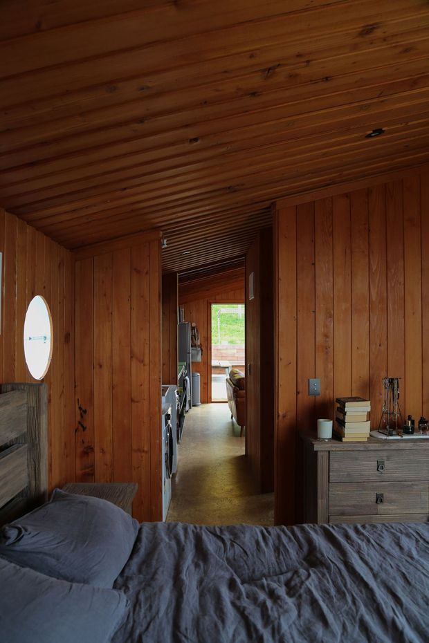 A compact, warmly lit bedroom clad throughout in natural timber boards, with a low timber-lined ceiling, a bed in the foreground, and a rustic wooden dresser. An open doorway leads through a narrow timber-lined hall toward a living area and glazed exterior door, while a round porthole-style window provides daylight on the left wall.
