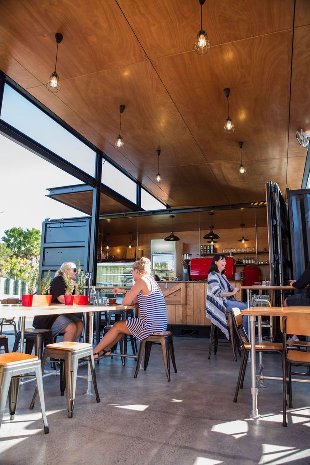 A bright, open café interior with a warm timber-panelled ceiling, exposed dark metal framing, large operable glazed openings, pendant lights, a timber service counter and open kitchen display. Café tables and stools fill the polished concrete floor, with customers seated throughout. The space has an industrial-inspired fit-out softened by warm wood and abundant daylight.