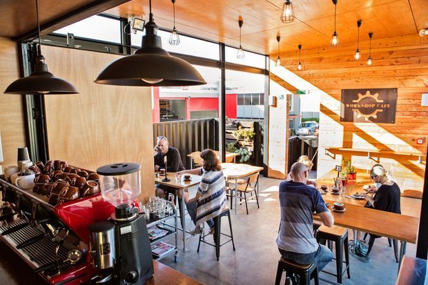 A bright, rustic-modern café interior with exposed timber ceiling and wall cladding, industrial pendant lights, polished concrete flooring, communal and individual timber tables, and a prominent coffee bar with espresso equipment. Large glazed openings connect the café to the street, while warm sunlight creates strong diagonal shadows across the timber wall.