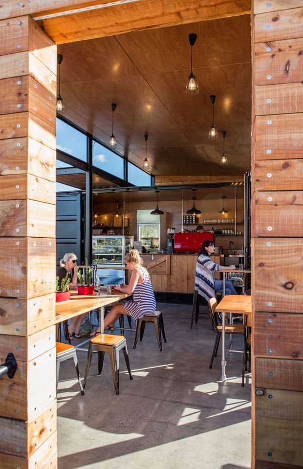 A bright café interior viewed through a wide timber-framed opening. The space features extensive warm timber wall and ceiling finishes, exposed dark steel framing, polished concrete flooring, pendant lights, a timber service counter with a red coffee machine, refrigerated display case, shelving, and casual tables with stools. Customers are seated in the café, with strong sunlight entering through large glazed openings.