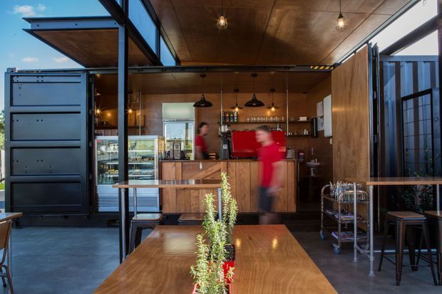 A compact contemporary café interior with an open service counter, red espresso machine, timber cabinetry and walls, pendant lighting, display refrigeration, and communal timber tables. Large glazed openings and dark steel framing connect the café to the outdoors, while commercial kitchen equipment and shelving are visible behind the counter.