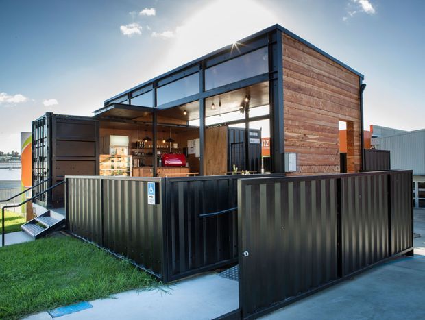 Exterior view of a compact contemporary modular commercial kiosk or container-based venue, clad in horizontal weathered timber and framed with dark metal. Large glazed openings and folding doors reveal a bar or service counter inside, while a black corrugated-metal perimeter enclosure forms an accessible deck or entry area. The building is set beside a paved commercial environment with some lawn and water visible in the background.