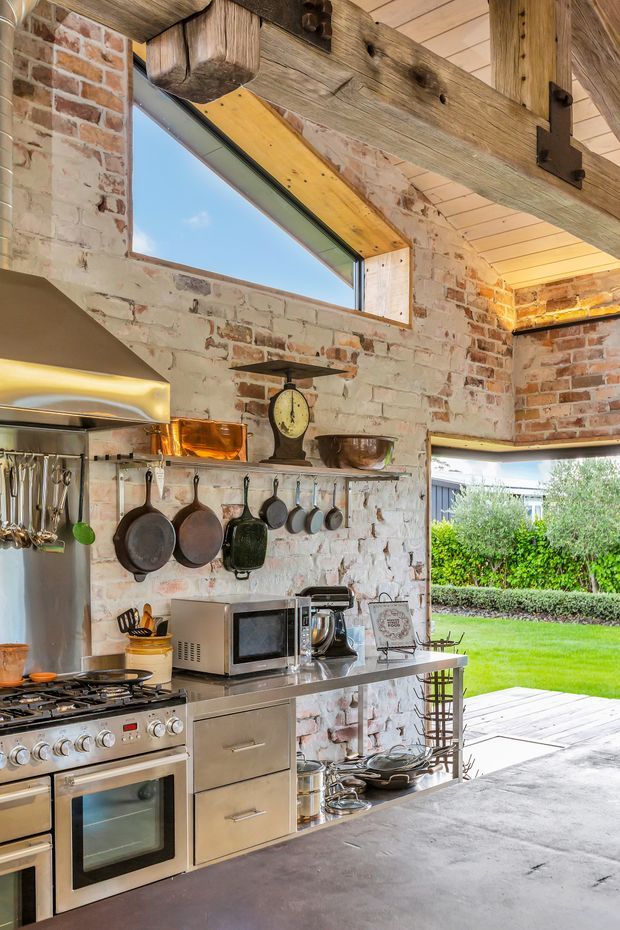 A rustic, industrial-style kitchen in a converted brick structure, featuring exposed aged brick walls, heavy reclaimed timber beams and ceiling boards, stainless-steel appliances and cabinetry, open shelving, hanging cookware, and a large stainless-steel range hood. A wide opening connects the kitchen to a timber deck and landscaped garden outside.