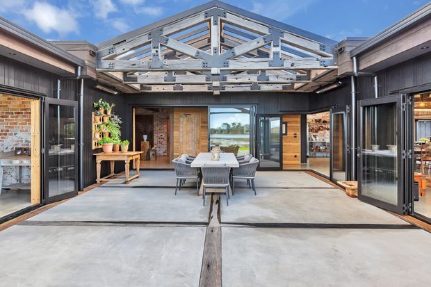 A contemporary covered courtyard or outdoor dining area framed by dark vertical cladding, exposed timber trusses and extensive glass folding doors. A central outdoor dining table and chairs sit on a concrete slab floor, with exposed brick walls, timber doors, built-in shelving and potted plants around the perimeter. The open roof structure reveals blue sky and a distant landscape beyond the glazing.