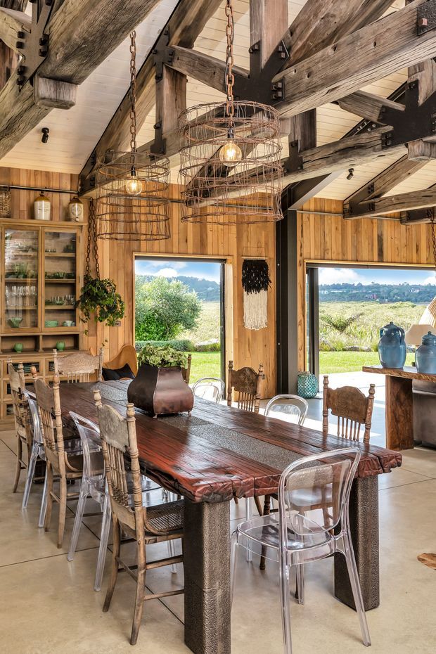 A rustic dining area with a long live-edge timber dining table surrounded by a mix of wooden and transparent acrylic chairs. The room features extensive exposed reclaimed timber beams, timber wall panelling, a pale pitched ceiling, wrought-metal cage pendant lights, open built-in shelving, and large windows overlooking a green rural landscape. The space blends rustic farmhouse character with industrial accents and abundant natural light.