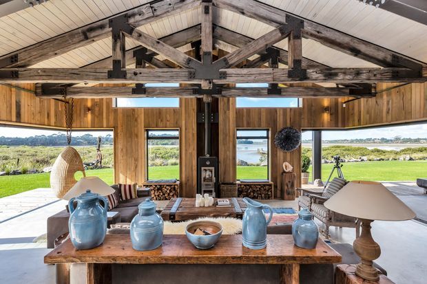 A rustic, open-sided living room or lounge with expansive glazing overlooking a grassy coastal or rural landscape and water beyond. The room features substantial exposed timber post-and-beam framing, a white pitched ceiling, vertical timber wall panelling, a central fireplace, and relaxed furnishings including woven seating, striped upholstered chairs, a timber console table, and blue ceramic vessels.