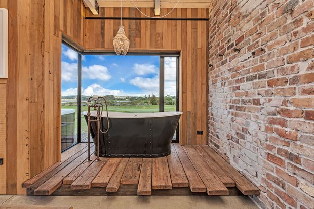 Rustic industrial-style bathroom featuring a freestanding dark bathtub on a raised timber platform. The room combines warm vertical timber wall cladding and timber flooring with an exposed red brick feature wall, exposed ceiling beams, and large corner glazing overlooking a green landscape. A woven pendant light adds a natural decorative element.