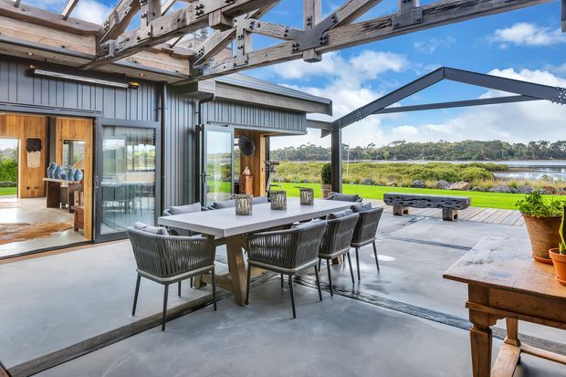 A spacious covered outdoor entertaining area adjoining a contemporary home, with a large dining table and woven chairs on a polished concrete floor. Timber-framed glazing and dark corrugated metal cladding define the house, while a timber pergola structure and steel canopy frame the open view toward a landscaped waterfront setting.