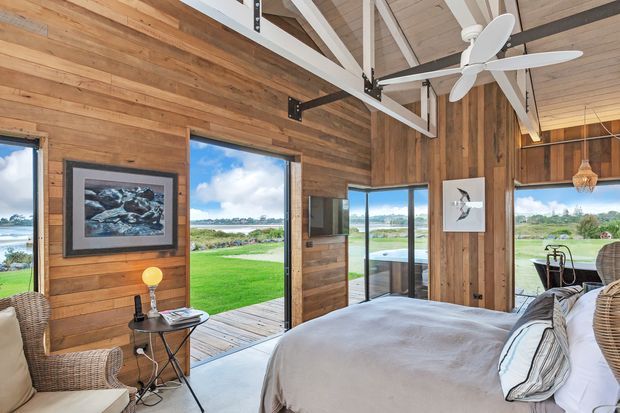 A bright, rustic bedroom with full-height glazing opening to a green waterfront landscape. The room features warm timber wall cladding and a timber-lined vaulted ceiling with exposed white-painted beams, a large white ceiling fan, woven wicker seating, and a neatly made bed. A small outdoor deck and pool or water feature are visible beyond the doors.