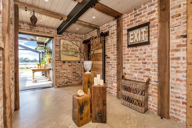 Rustic industrial-style entry hall with exposed red brick walls, substantial timber posts and beams, a light timber-panelled ceiling, and a polished concrete floor. Decorative timber pedestal tables, a framed artwork, woven basket, and prominent EXIT sign furnish the space. An open glazed doorway leads to a bright exterior area.