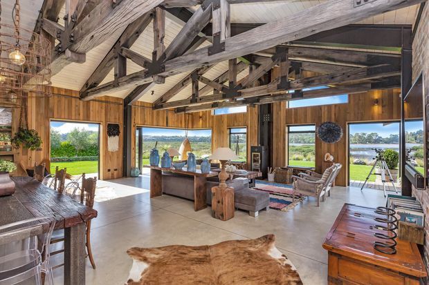Open-plan rustic living and dining interior with exposed heavy timber trusses, warm vertical timber wall panelling, a concrete floor, and extensive windows overlooking a green rural landscape and water. The room includes a large timber dining table, lounge seating, a central console, a large hide rug, and vintage-style furnishings.