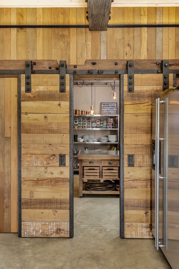 A rustic industrial kitchen or pantry interior featuring large reclaimed timber sliding barn doors with dark metal hardware. The doors open onto a compact, neatly organised pantry with open shelving, labelled storage containers, dishes, drawers and a utility sink. Vertical timber wall panelling, exposed dark timber and metal structural elements, pendant lighting, stainless-steel refrigeration and a polished concrete floor reinforce the utilitarian character.