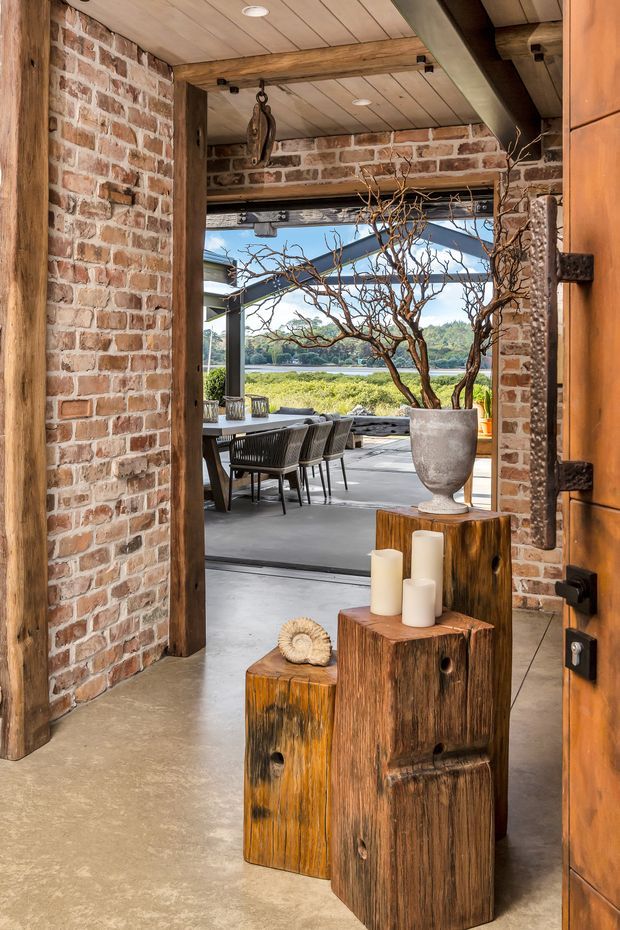 A rustic, industrial-style entryway framed by exposed red brick walls, heavy timber beams, and a timber-lined ceiling. The open doorway leads to an outdoor dining terrace with a table and chairs, while large timber pedestal blocks, candles, and a sculptural branch arrangement create a warm natural focal point. A broad glazed opening provides views toward water and landscaped surroundings.