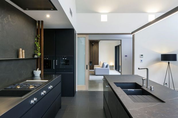 Contemporary open-plan kitchen with dark matte cabinetry, a large stone island and integrated cooktop, sink, and tapware. The space features dark tiled flooring, a charcoal textured wall, concealed appliances, linear lighting, and a broad opening toward a bright living area with timber screening.