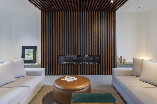 Contemporary living room with two pale upholstered sofas facing a round brown leather ottoman and a glass coffee table. A dramatic floor-to-ceiling dark timber slatted feature wall incorporates a linear fireplace and continues across the ceiling, contrasting with white walls and a neutral carpet. Recessed ceiling lighting and restrained furnishings create a refined, warm interior.