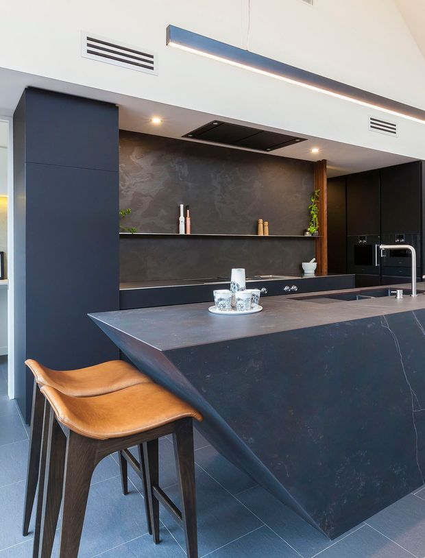 A contemporary kitchen featuring a large dark stone island with an angular waterfall edge, integrated dark cabinetry, a matching textured dark splashback, black appliances and fixtures, and open shelving. Two tan leather bar stools sit at the island. Linear pendant lighting, recessed spotlights, and white ceilings provide contrast to the predominantly charcoal fit-out.