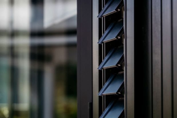 Close-up architectural detail of a dark vertical louvred facade or window assembly. Blue-grey angled metal louvres are set within a narrow opening beside dark cladding, with blurred glazed building surfaces in the background.