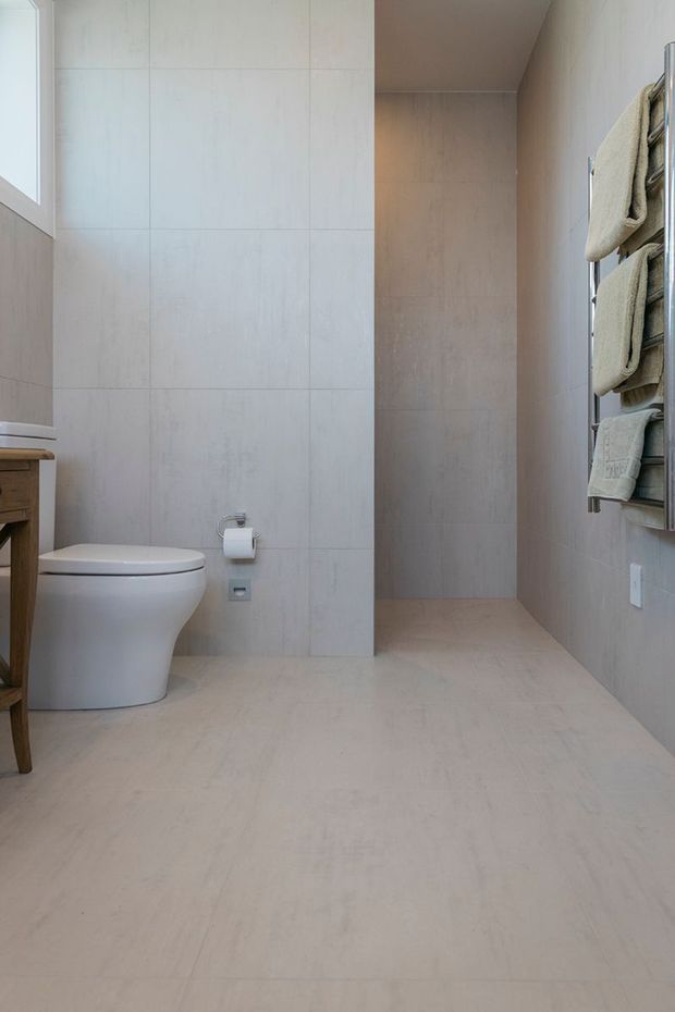 A bright, minimalist bathroom with large pale beige stone-look tiles covering the walls and floor. The room includes a white wall-hung toilet, a partially visible timber vanity or shelving unit, a small window, and a wall-mounted heated towel rail holding neutral towels. A recessed shower area is visible at the rear.