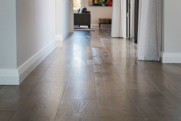 A bright residential hallway viewed at floor level, featuring wide-plank timber flooring in a medium grey-brown tone, white walls and trim, and doorways with light curtains leading toward a living area.