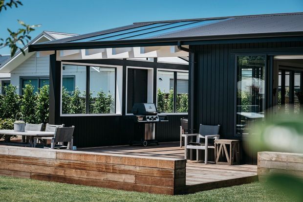Contemporary single-storey home viewed from the garden, featuring a dark vertical-clad outdoor entertaining pavilion with a broad timber deck, built-in outdoor dining area, barbecue, and large glazed openings. A lighter main house with a pitched metal roof is visible behind, with landscaped screening and raised timber garden edging in the foreground.