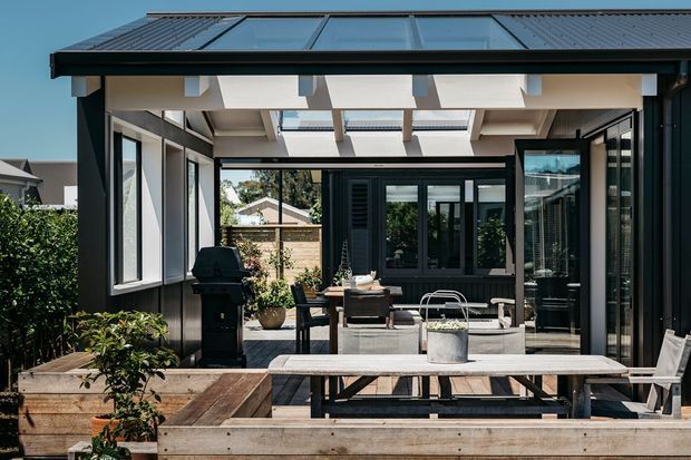 A modern enclosed outdoor living and dining area with dark metal-framed glazing, a translucent roof canopy, exposed light-coloured beams, and timber planter boxes. The space includes a long dining table, outdoor seating, a barbecue, and a paved courtyard-style deck, with neighbouring residential buildings visible beyond.