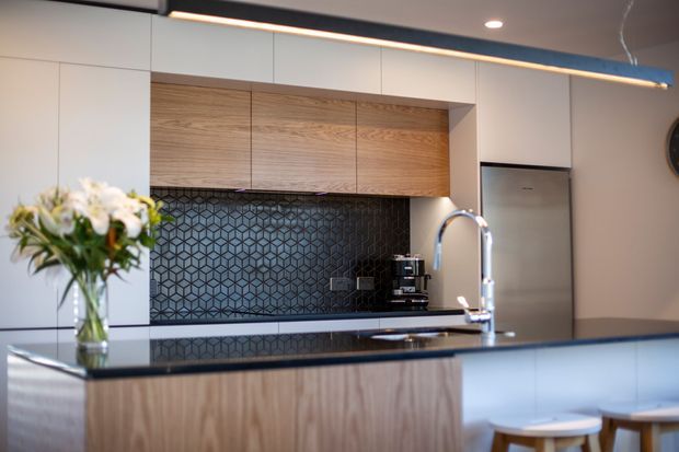 A contemporary kitchen with white full-height cabinetry, warm timber overhead and island joinery, a dark geometric patterned splashback, dark benchtops, stainless-steel appliances, and a curved chrome tap. Timber stools sit at the island, while a slim linear pendant provides accent lighting.
