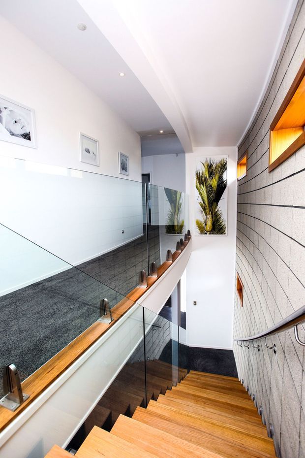 Bright contemporary stairway viewed from above, with warm timber treads, clear glass balustrades, a dark carpeted landing, white walls and ceiling, and a textured grey masonry feature wall. A timber handrail and recessed illuminated wall niches add warmth, while indoor plants and framed artwork are visible along the upper landing.